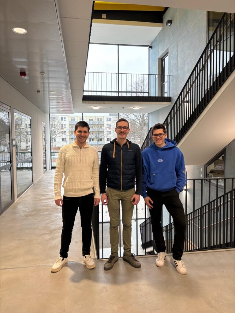 Drei Männer in einem modernen Treppenhausflur, die nebeneinander stehen und in die Kamera lächeln. Links: ein Mann mit kurzen dunklen Haaren, cremefarbener Strickpullover mit Muster, schwarze Hose, weiße Sneakers; die Arme hängen locker. Mitte: ein Mann mit Brille und kurzem Haar, dunkelblaue Steppjacke mit hellem Reißverschluss/Details, olivgrüne Hose, dunkle Schuhe; steht gerade, Hände locker neben dem Körper. Rechts: ein Mann mit Brille, blauem Hoodie, schwarze Hose, weiße Sneakers; er lehnt lässig am Geländer und hat ein Bein angewinkelt.